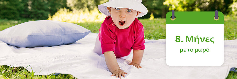 Ένα μωρό με κόκκινο πουκάμισο και λευκό καπέλο σέρνεται πάνω σε μια λευκή κουβέρτα σε μια χορταριασμένη περιοχή, με ένα γκρίζο μαξιλάρι κοντά του.