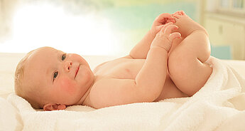 Ένα μωρό ξαπλώνει σε μια λευκή κουβέρτα, κρατώντας το πόδι του.