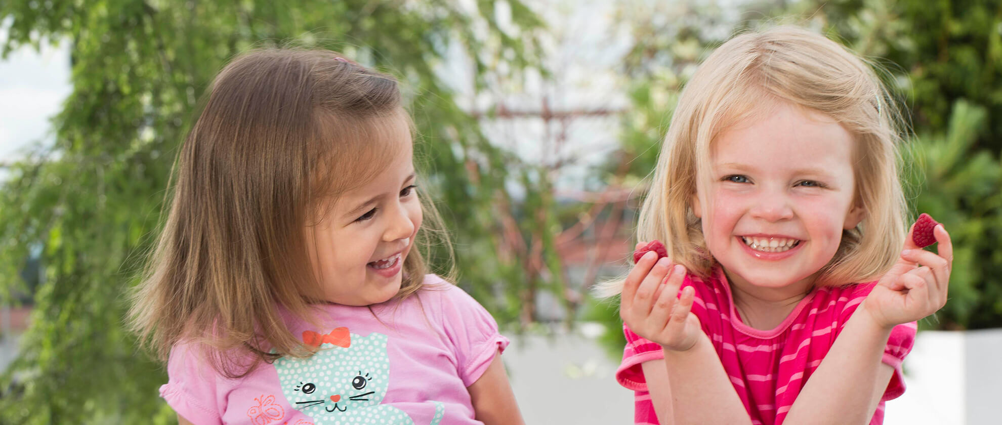 Two young girls, one in a pink shirt with a cat design and the other in a striped pink shirt, are holding and looking at red raspberries outdoors.