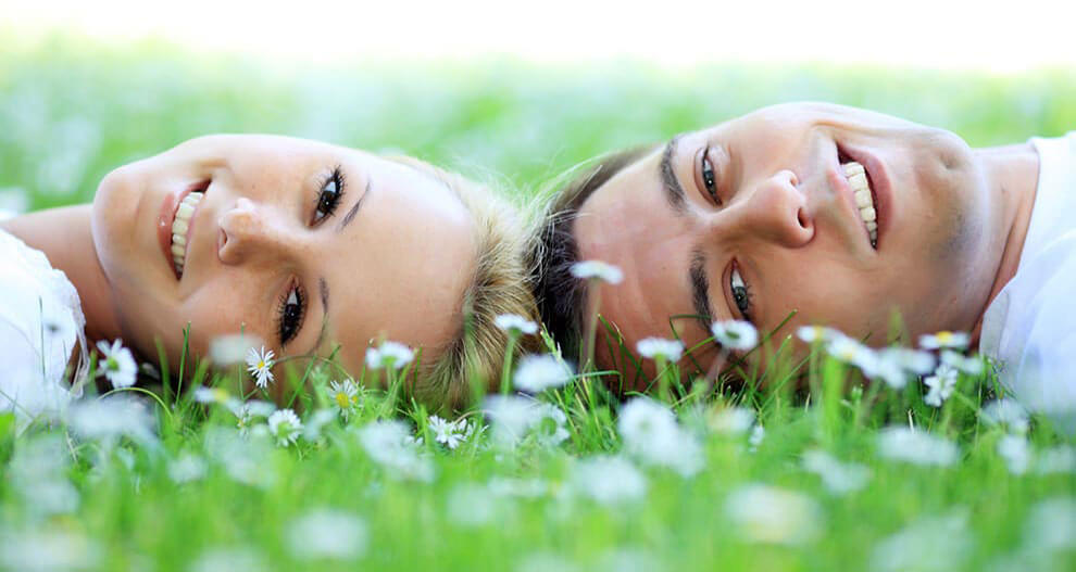Two individuals lie on a grassy field surrounded by white flowers, looking directly at the camera.