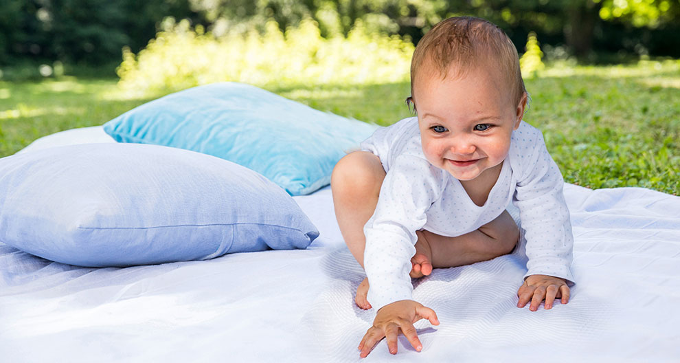 A baby in a white outfit is crawling on a white blanket in a grassy area with a blue pillow nearby.
