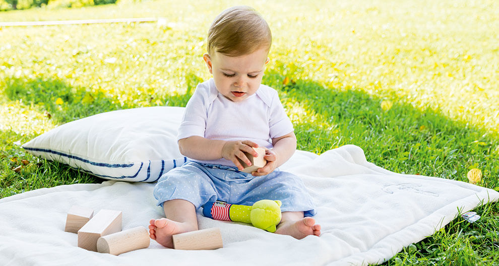 A baby sits on a blanket outdoors, playing with wooden blocks and a toy.