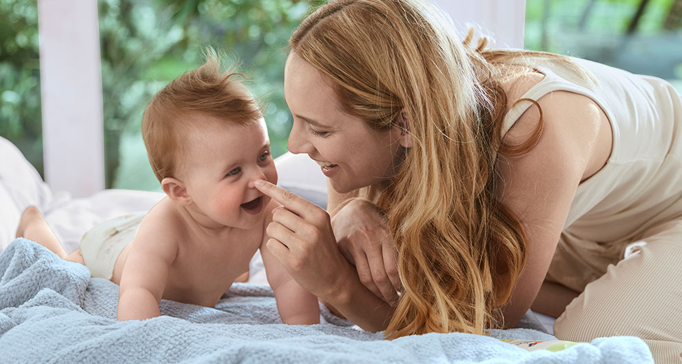 A woman and a baby are lying on a bed, with the woman touching the baby's nose.