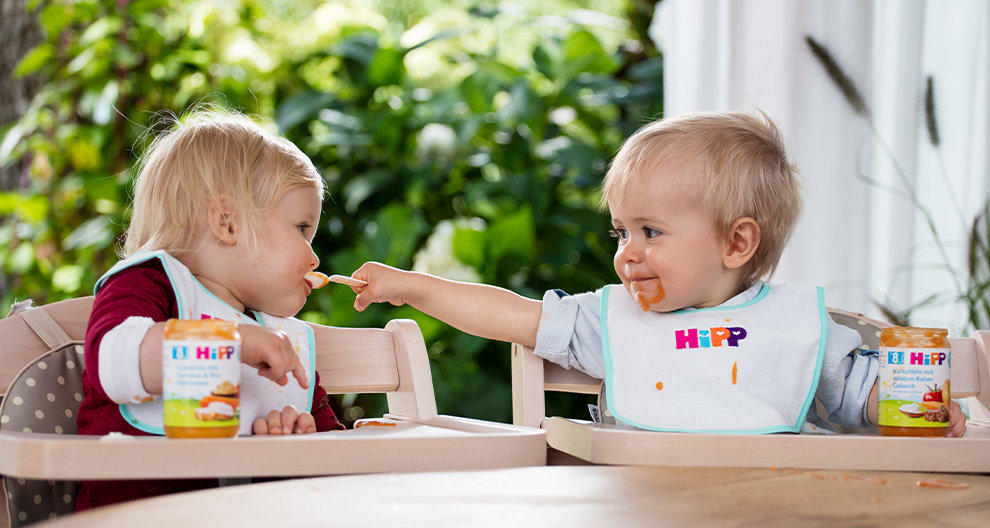 Two toddlers are seated at a table, feeding each other with food from a jar.