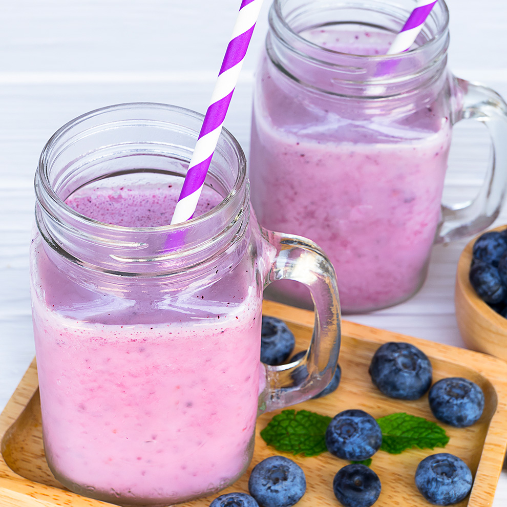 Two glass jars filled with a pinkish smoothie, each with a purple and white striped straw, sit on a wooden tray. Scattered around the tray are several blueberries and a sprig of mint.