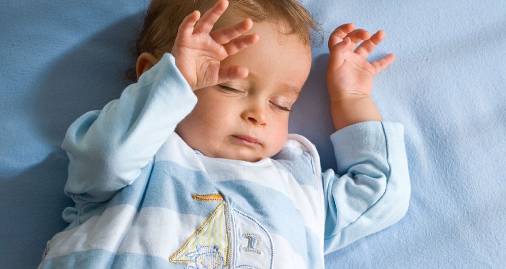A baby lies on a blue surface, wearing a light blue outfit with a sailboat design.