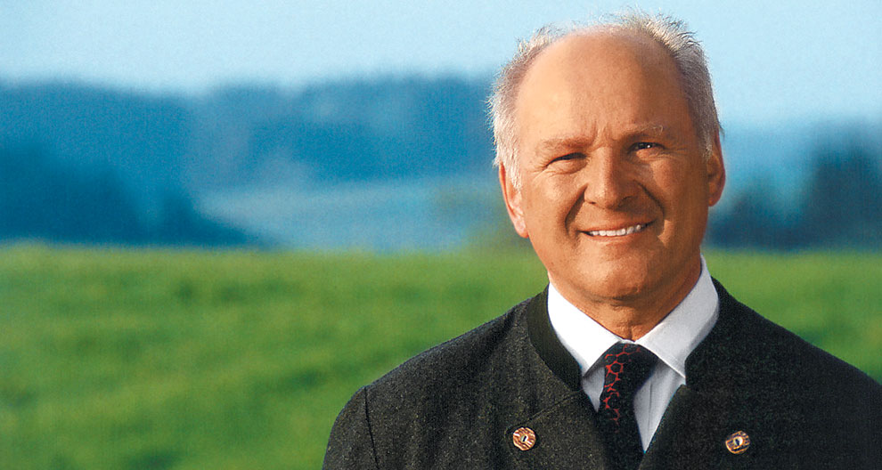 An elderly man in a suit stands against a scenic backdrop of a field and distant hills.