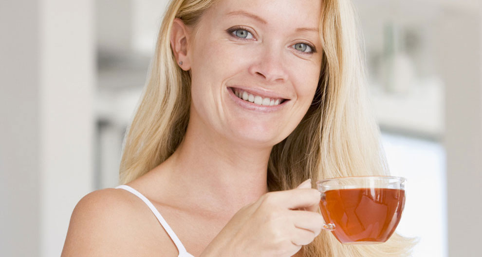 A woman with blonde hair and blue eyes holds a glass of amber-colored liquid.