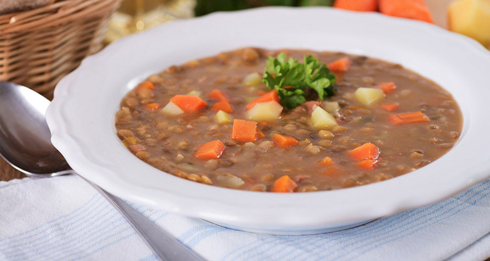 A bowl of thick, hearty soup with vegetables and grains is presented on a table with a spoon and a basket of bread.