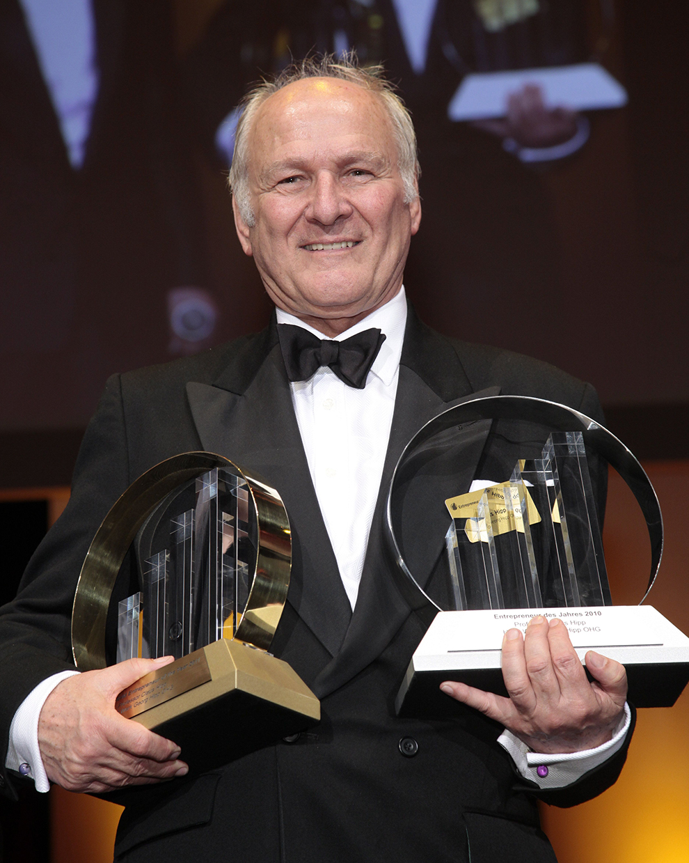 An elderly man in a black tuxedo holds two awards, one with a gold rim and the other with a silver rim, both featuring a clear crystal structure.