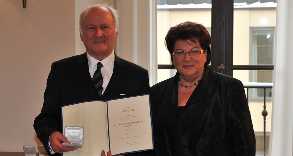 An elderly man in a black suit holds a certificate and a round silver object, standing next to an elderly woman in a black outfit, both posing for a photograph in a room with a window and a wall-mounted light fixture.